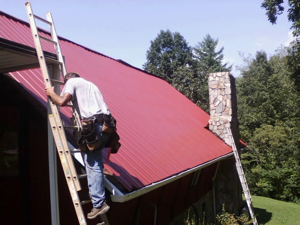 Roofer on a ladder beside a red metal roof and stone chimney — Soffit & Fascia Repair by Roofing Brothers in Gentry